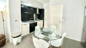 A compact kitchen with a glass dining table and white chairs, a small stool, and a white cabinet. - Cardeñosa 1 / Asamblea Centro (Madrid)