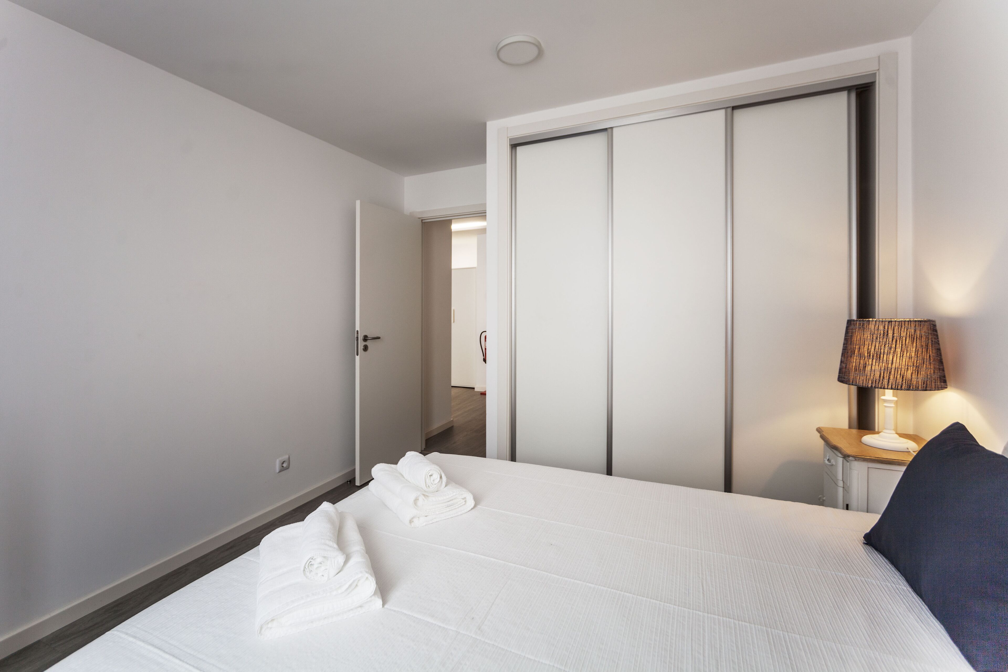 A hotel room with a bed, a nightstand with a lamp, a door, and a view of another room.