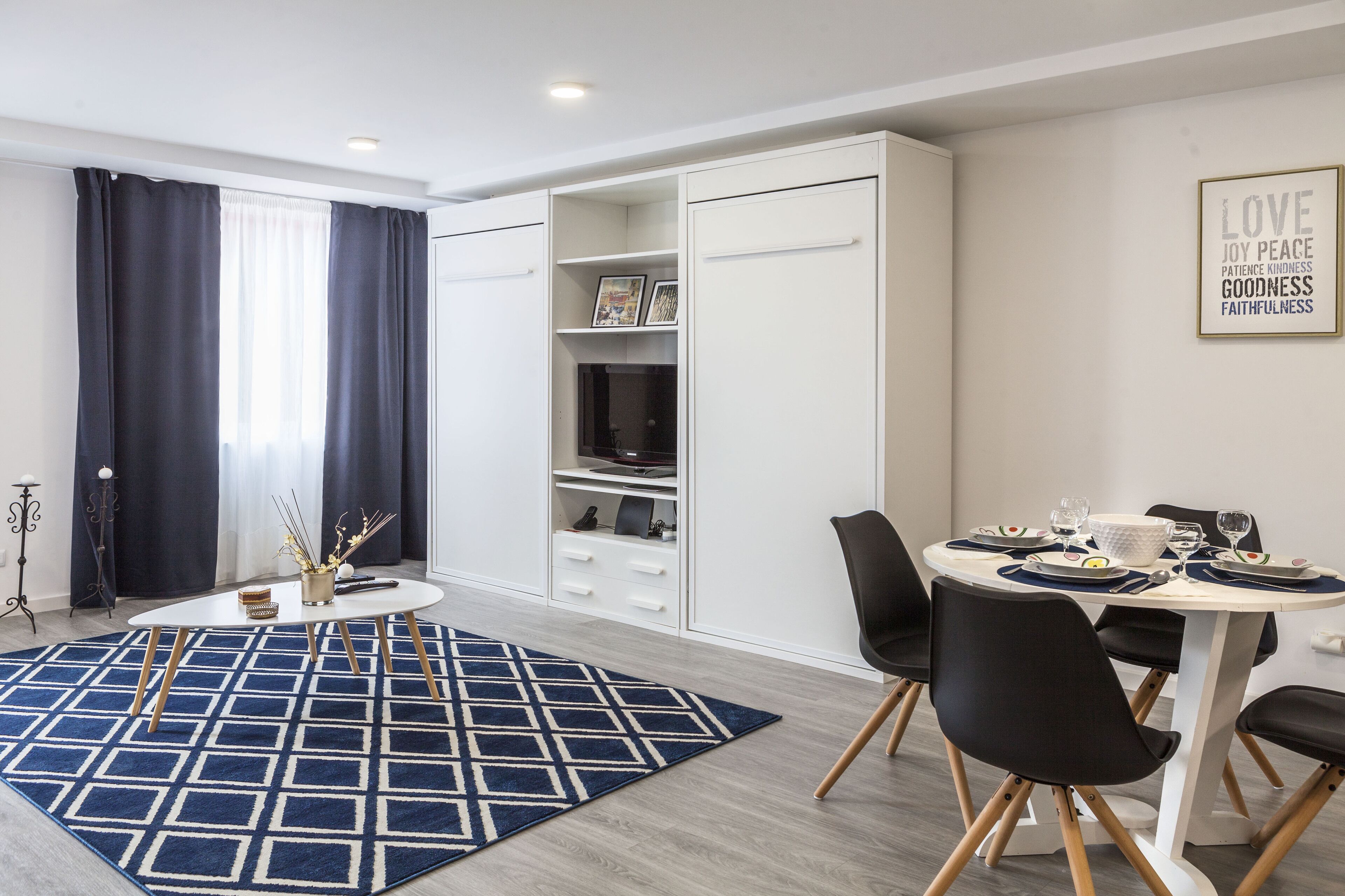 A modern living room with a dining area, featuring a white cabinet with open shelves, a round dining table set for four, and a blue patterned rug.