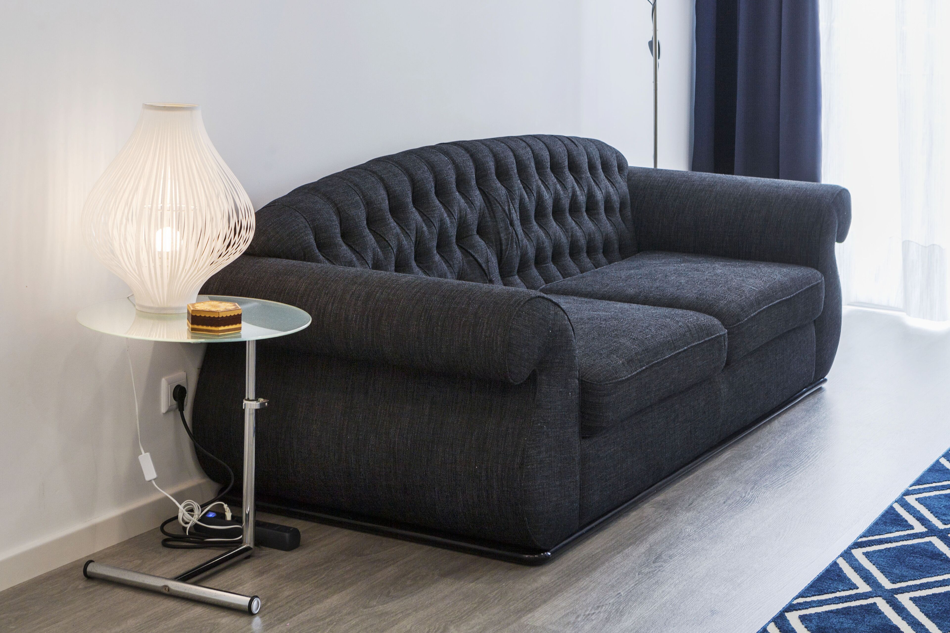 A dark grey sofa with a tufted backrest, a modern white lamp with a ribbed design, and a small wooden object on a glass table beside it.