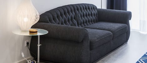 A dark grey sofa with a tufted backrest, a modern white lamp with a ribbed design, and a small wooden object on a glass table beside it.