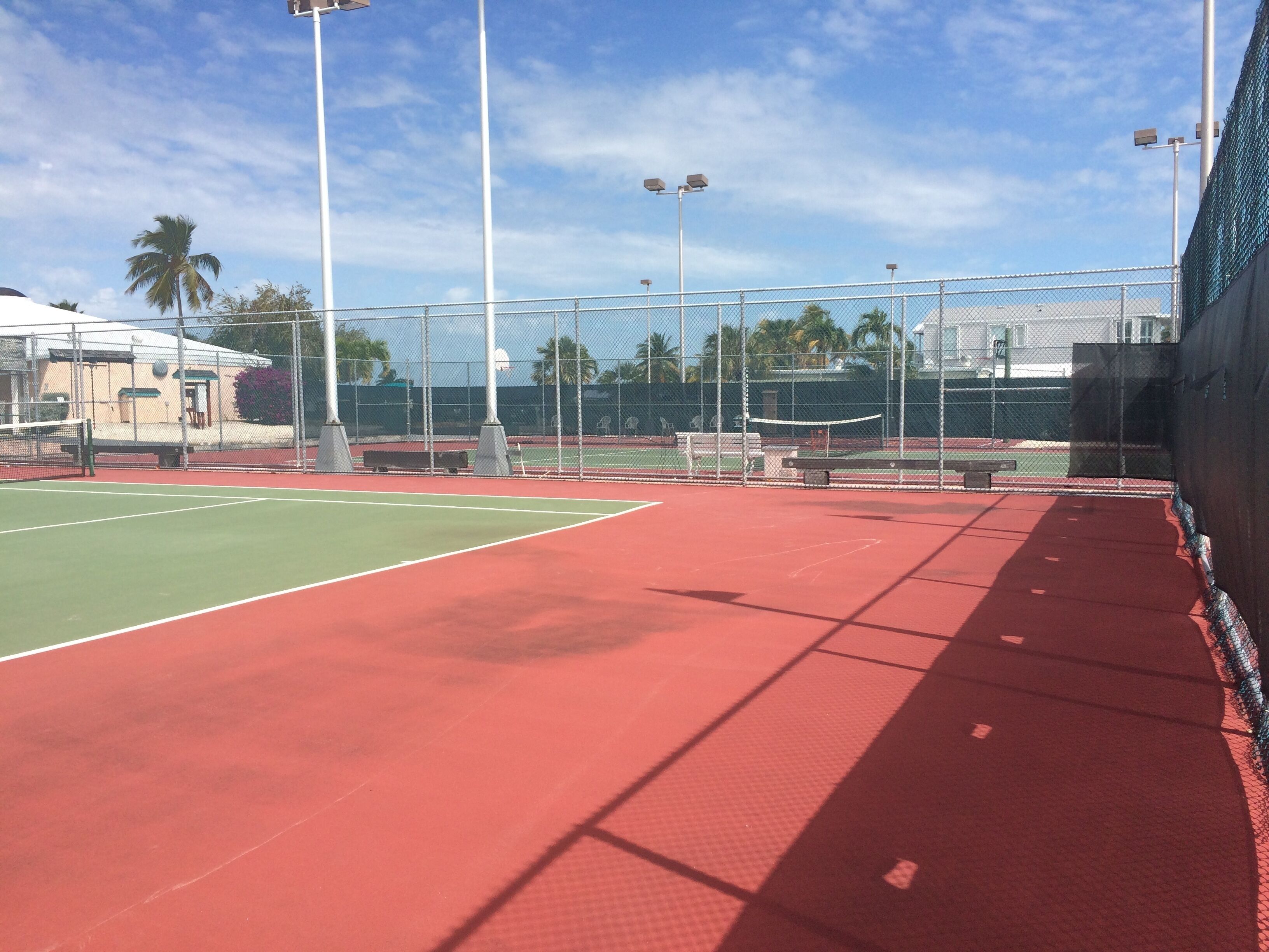 An outdoor tennis court with a red surface, surrounded by a chain-link fence, and equipped with floodlights.