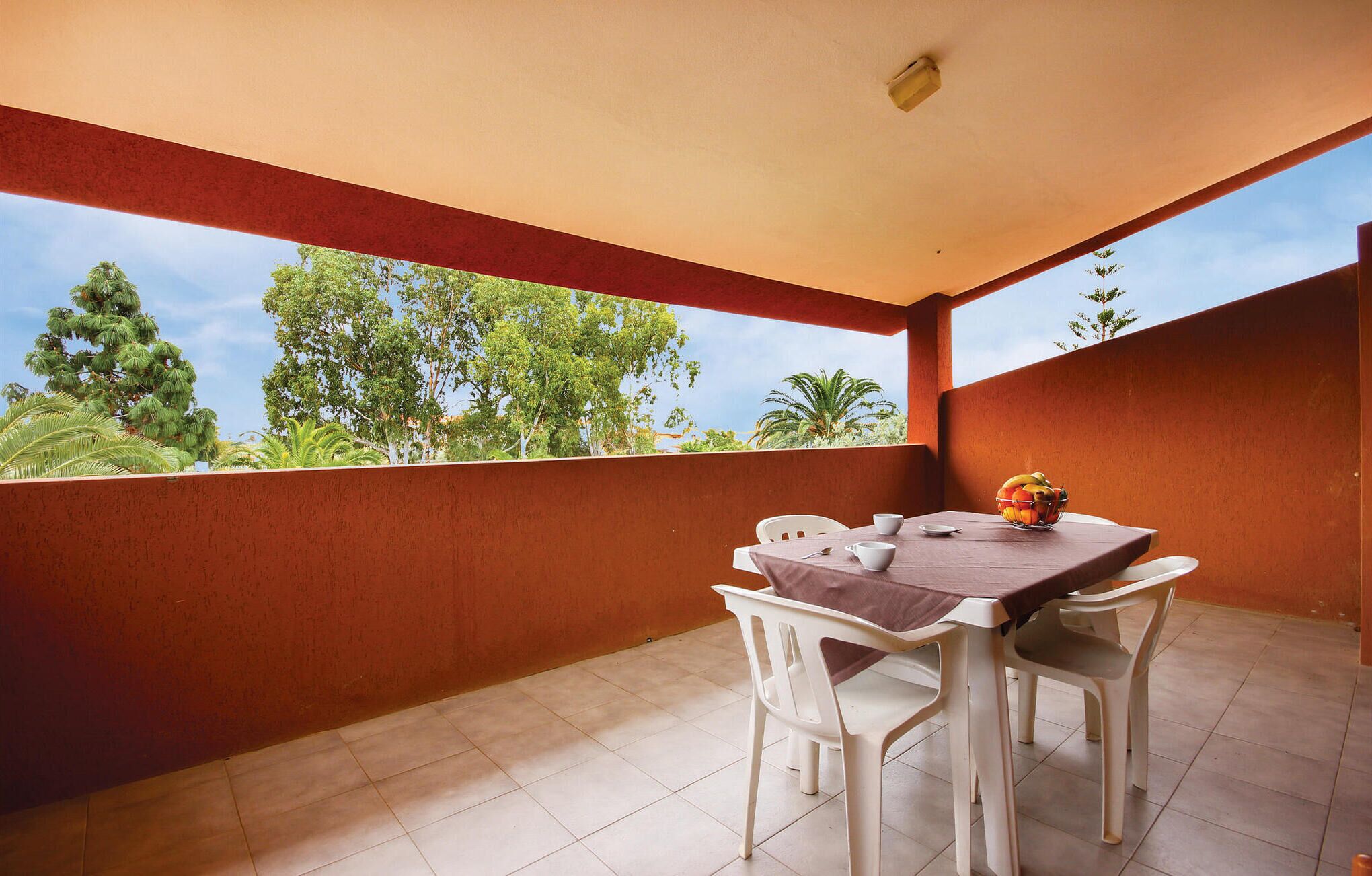 A covered outdoor dining area with a table and chairs, a bowl of fruit, and a view of trees and sky.