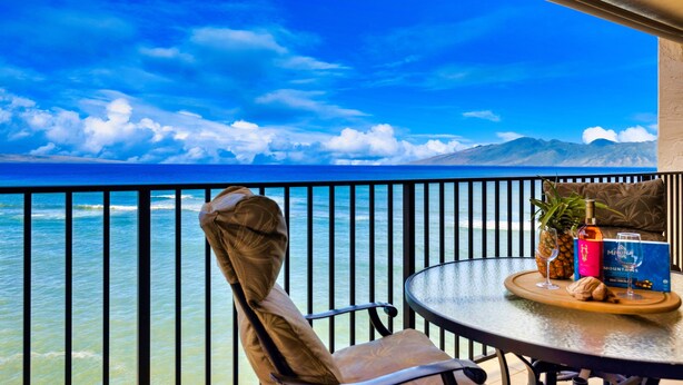 A balcony with a view of the ocean and mountains, a chair, a table with a tray of food and drinks, and a bottle of wine.