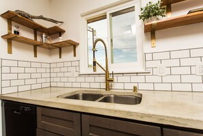 Sink with gold faucet and white tiled backsplash, accompanied by wooden shelves and a window with outdoor view.