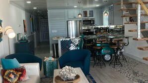Ein geräumiges Wohnzimmer mit einem weißen Sofa, blauen Sesseln und einem Couchtisch mit dekorativem Mittelstück. Im Hintergrund ist eine Küche mit modernen Geräten und ein Essbereich mit einem Holztisch und Stühlen zu sehen.
