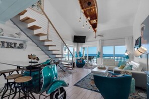 A spacious living area with a large window overlooking the sea, a teal vintage scooter, a staircase, and a cozy seating area with a white sofa and blue armchairs. - SPECTACULAR, TOTALLY RENOVATED 3 BEDROOM + CONDO ON BEACH, GORGEOUS GULF VIEWS  (Miramar Beach)