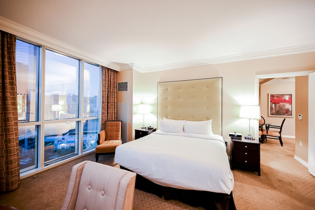 A hotel room with a large bed, a chair, a desk, and a view of the city through large windows.