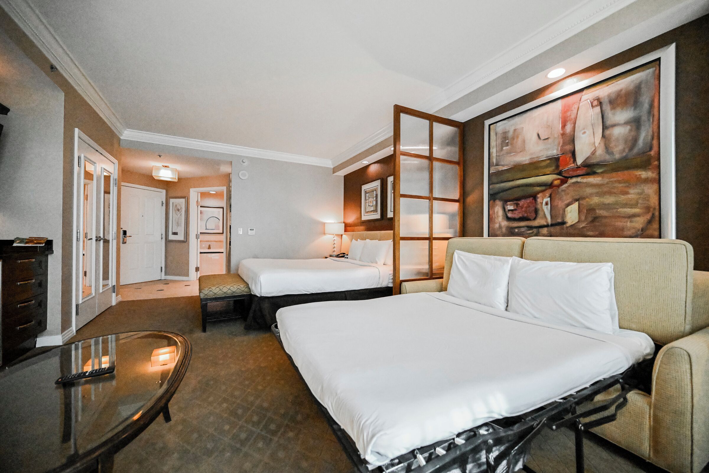 A hotel room with two beds, a sofa, a glass table, and a painting on the wall.