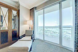 Quarto de hotel com uma grande porta de vidro deslizante para uma varanda, um banco estofado e um abajur.