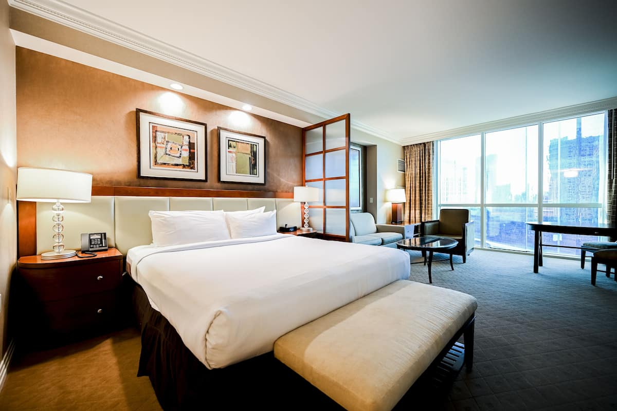 A hotel room with a large bed, a desk, a chair, and a view of the city through large windows.