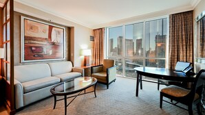 A hotel room with a large window, a sofa, a coffee table, a desk with a laptop, and a painting on the wall. - StripViewSuites Two Bedroom at Signature (Las Vegas)