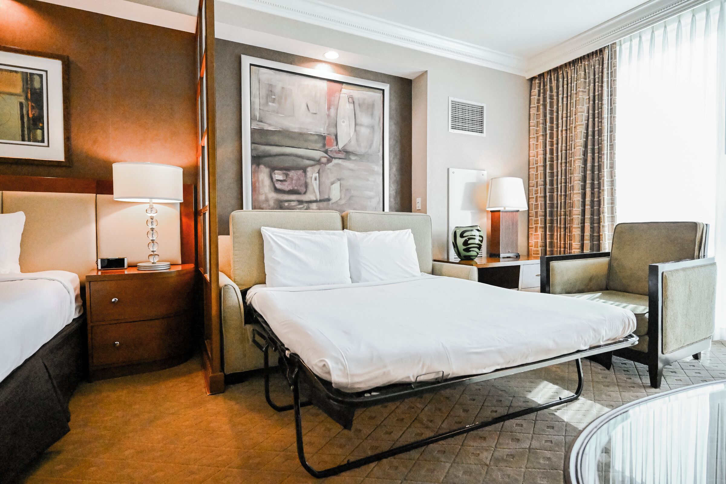 A hotel room with a bed, bedside tables, a desk, a chair, and a large abstract painting on the wall.