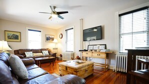 A living room with a brown leather sofa, a wooden coffee table, a wooden chest, a television on a stand, and a ceiling fan. - Atlantic Street Guesthouse - The Best Kept Secret! (Newport)