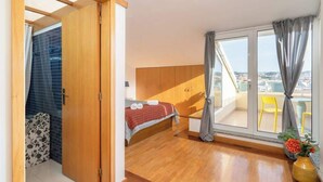 A bedroom with a bed, a window with curtains, a wooden wardrobe, and a view of the city. - HostWise - Seaside Beach Flat (Vila Nova de Gaia)