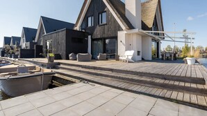 Ein modernes Haus mit einer Holzterrasse, ein Boot liegt in der Nähe im Hafen, und es gibt einen Blick auf das Wasser.
