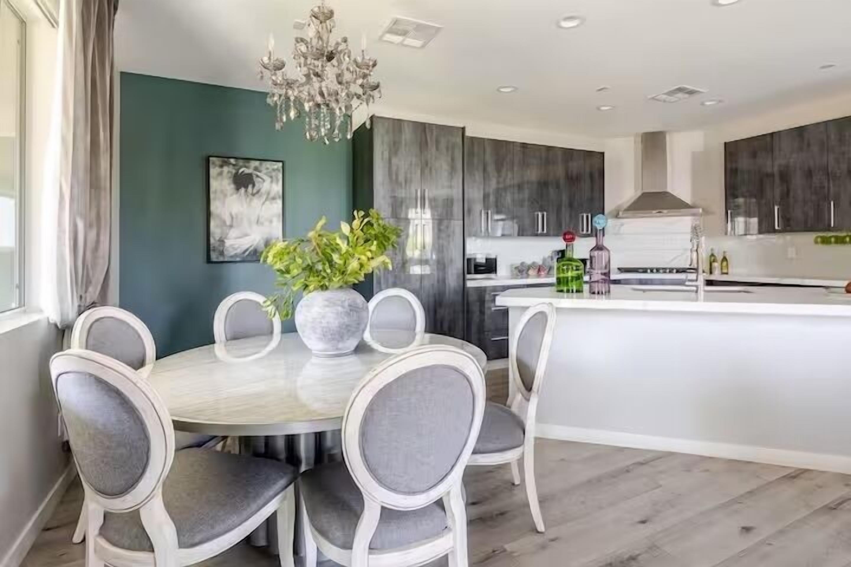A modern kitchen with a central island, grey upholstered chairs, a chandelier, and a green accent wall.