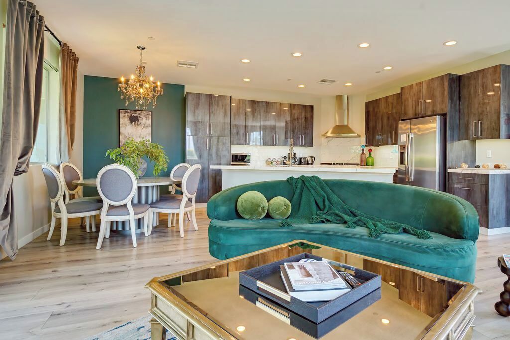 A modern living room with a green sofa, dining table, and chairs. A kitchen with dark cabinets and a refrigerator is visible in the background.