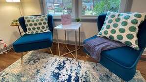 Una habitación con dos sillones azules, cada uno con un cojín verde de lunares, una mesita blanca con una caja rosa que dice 'Relax', y una ventana que da a un barrio residencial.