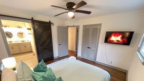 Un dormitorio con cama, almohadas, ventilador de techo, televisor que muestra una chimenea, y un baño visible al fondo.