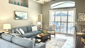 A spacious living room with a grey sofa, a coffee table, and a large window with blinds.