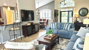 A spacious living room with a large flat-screen TV, a comfortable sofa set, a coffee table, and a dining area with a chandelier.