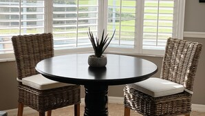 A round table with a potted plant in the center, flanked by two wicker chairs with white cushions. Large windows with horizontal blinds provide a view of a green outdoor area.