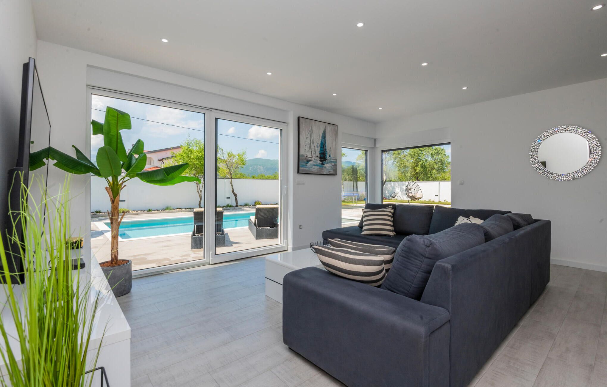 Ein modernes Wohnzimmer mit grauem Sofa, großem Fenster und Blick auf einen Pool.