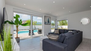 A modern living room with a grey sofa, a large window, and a pool view. - Lovely home in Donji Prolozac (Donji Prolozac)