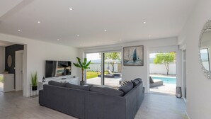 A modern living room with a grey sofa, a flat-screen TV, and a large window overlooking a pool and garden. - Lovely home in Donji Prolozac (Donji Prolozac)