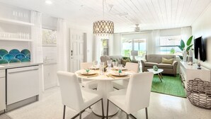 A modern kitchen with white cabinetry, a dining area with a round table and white chairs, a sofa, and a TV on the wall. - Sailfish Gulf Suites #1, Beachfront, Steps to the Sand (Holmes Beach)