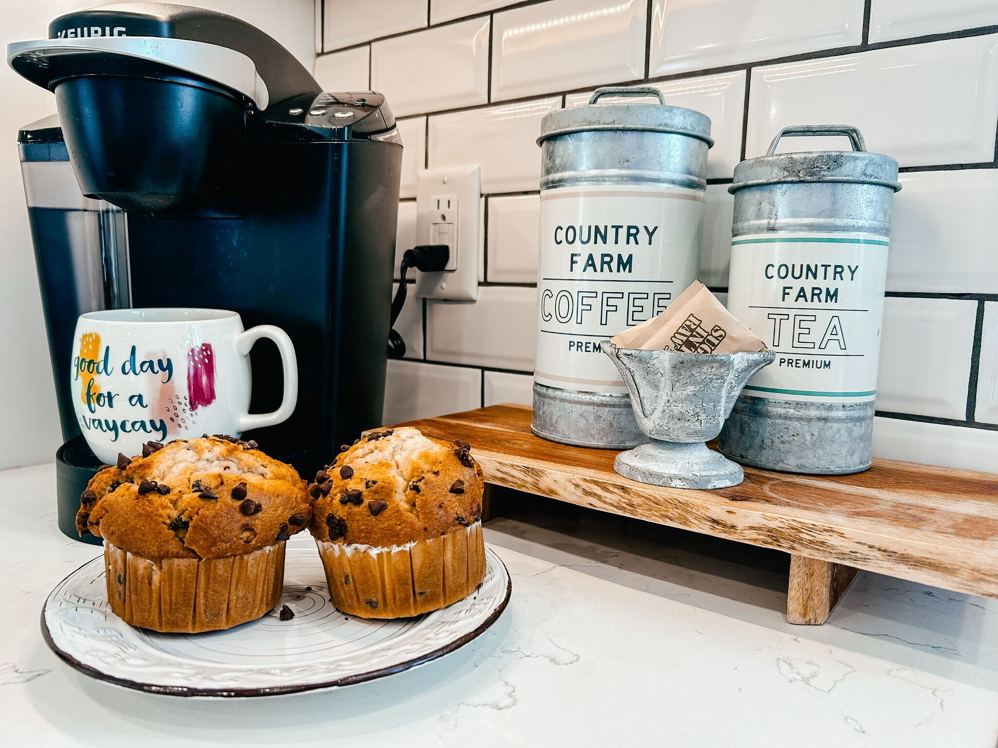 Una encimera de cocina con una cafetera, dos muffins y dos recipientes metálicos etiquetados como 'Country Farm Coffee' y 'Country Farm Tea'.