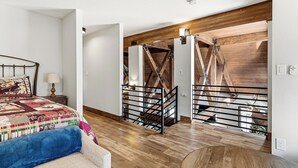 A bedroom with a bed, a nightstand, a lamp, a wooden floor, and a staircase with a metal railing.