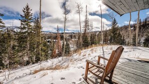 A wooden deck with a single wooden chair, overlooking a snowy landscape with trees and a clear sky.