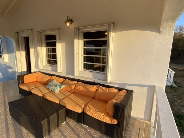 Un área de estar al aire libre techada, con un sofá y reposapiés de mimbre, ventanas amplias y una lámpara de pared.