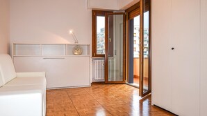 Una habitación con suelo de madera, un sofá blanco, un reloj montado en la pared y un ventanal corredero que da a un balcón al aire libre.
