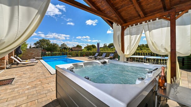 Un jacuzzi bajo un porche de madera con una piscina y sillas de descanso al fondo.