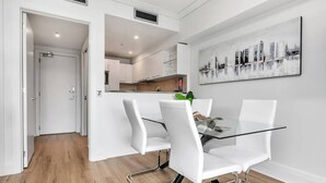 Un espace repas moderne avec une table en verre et des chaises blanches, un tableau représentant un paysage urbain accroché au mur, et une cuisine en arrière-plan.