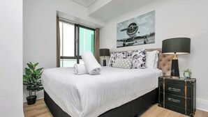 Une chambre à coucher avec un lit, deux lampes de chevet, une plante en pot, une valise et un tableau accroché au mur.
