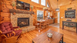 A cozy log cabin interior with a stone fireplace, wooden dining table, and a television mounted above it. - Breathtaking Views – No-Stair Entry, Hot Tub, Pool Table & Seasonal Pool Access (Sevierville)