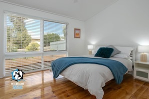 A bedroom with a large window, a bed with white bedding and a blue throw, two bedside tables with lamps, and a framed picture on the wall.