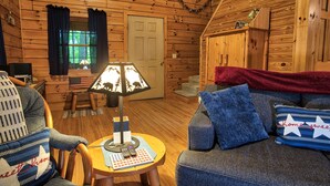 A cozy cabin interior with wooden walls, a sofa with blue cushions, a lamp, and a small table with a remote control and a book. - Beechwood Cabin near Ash Cave (New Plymouth)
