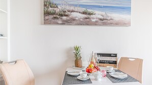 Una mesa de comedor con una piña, frutas, una botella y vasos, frente a un cuadro con temática playera.