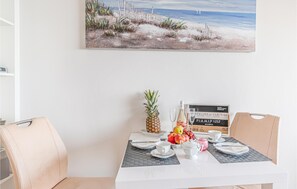 Ein Esstisch mit einer Ananas, Früchten, einer Flasche und Gläsern, vor einer Strand-Szene als Gemälde.