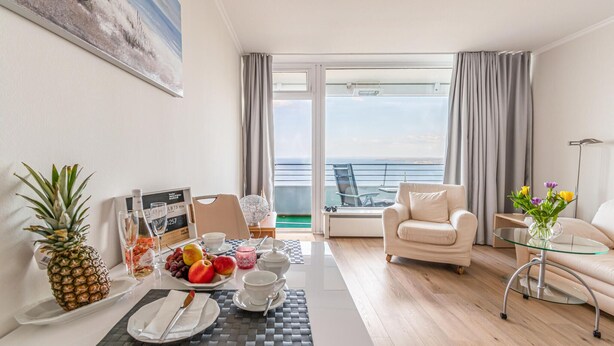 Una habitación de hotel moderna con balcón, un área de comedor con fruta y un menú, un sofá y una mesa de centro de vidrio.