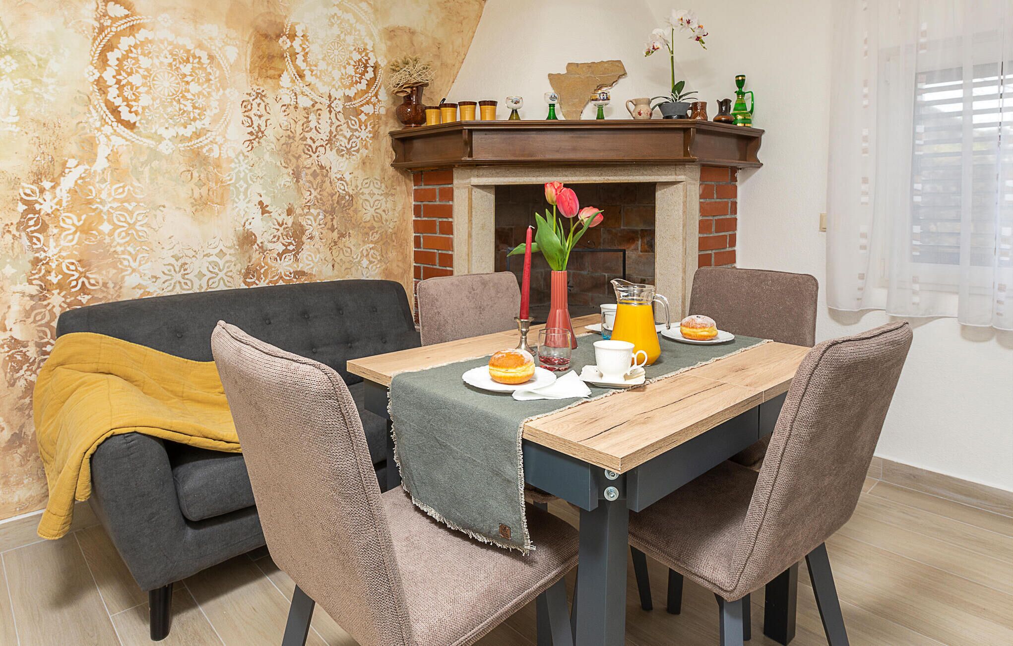A dining area with a wooden table, four chairs, a vase with flowers, a red candle, and a brick fireplace.