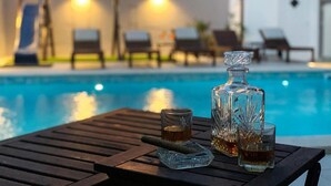 Taula al costat de la piscina amb ampolla i gots, una piscina i gandules al fons.