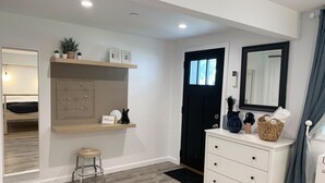 Una habitación moderna con piso de madera, un aparador blanco con cajones, una puerta negra, un espejo y una estantería con plantas y un letrero de bienvenida.