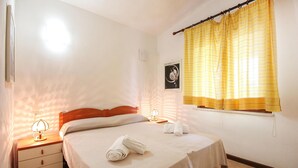 Een net opgemaakt bed met een opgevouwen handdoek, een bedlampje en een raam met gele gordijnen.