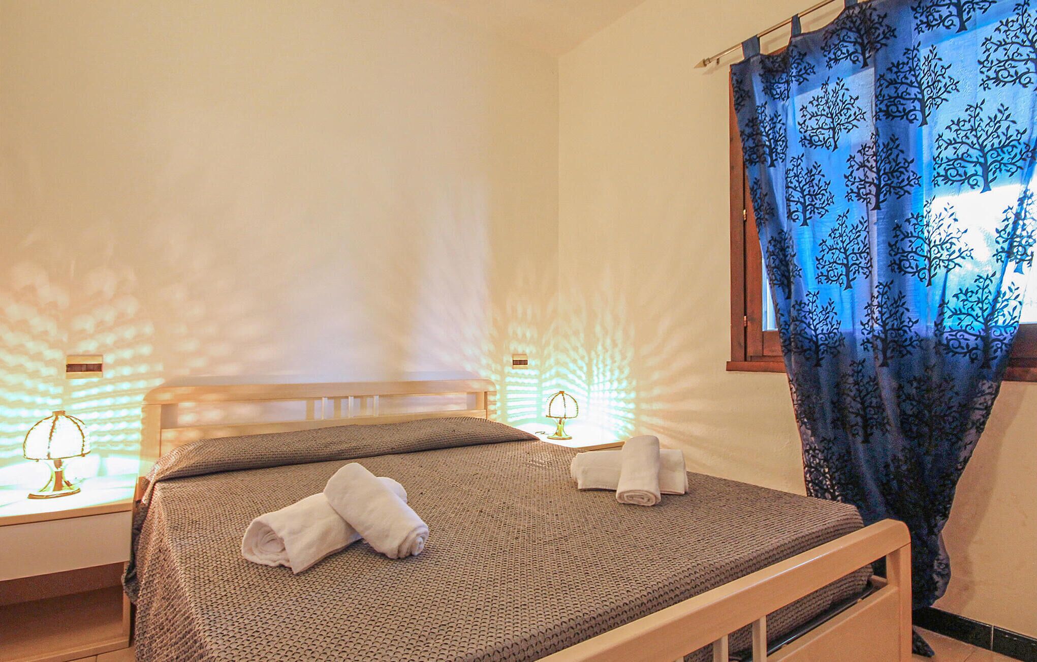 Un lit bien fait, avec deux serviettes enroulées, une lampe de chevet et une fenêtre ornée d’un rideau bleu.